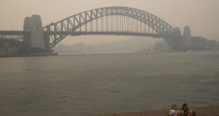 Two girls sit on a bench overlooking Sydney Harbour Bridge, which is shrouded in smoke haze from bushfires.
